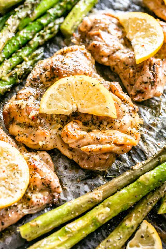 Close up overhead view of lemon garlic chicken thighs garnished with lemon slices, next to asparagus on a baking pan.