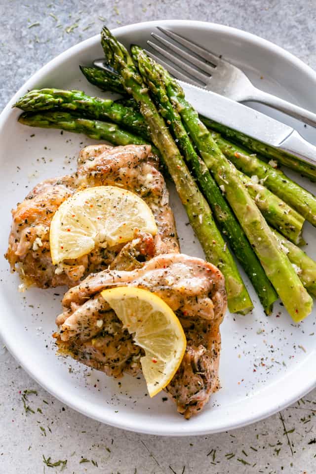 Overhead view of two lemon garlic chicken thighs garnished with lemon slices next to asparagus on a white plate.