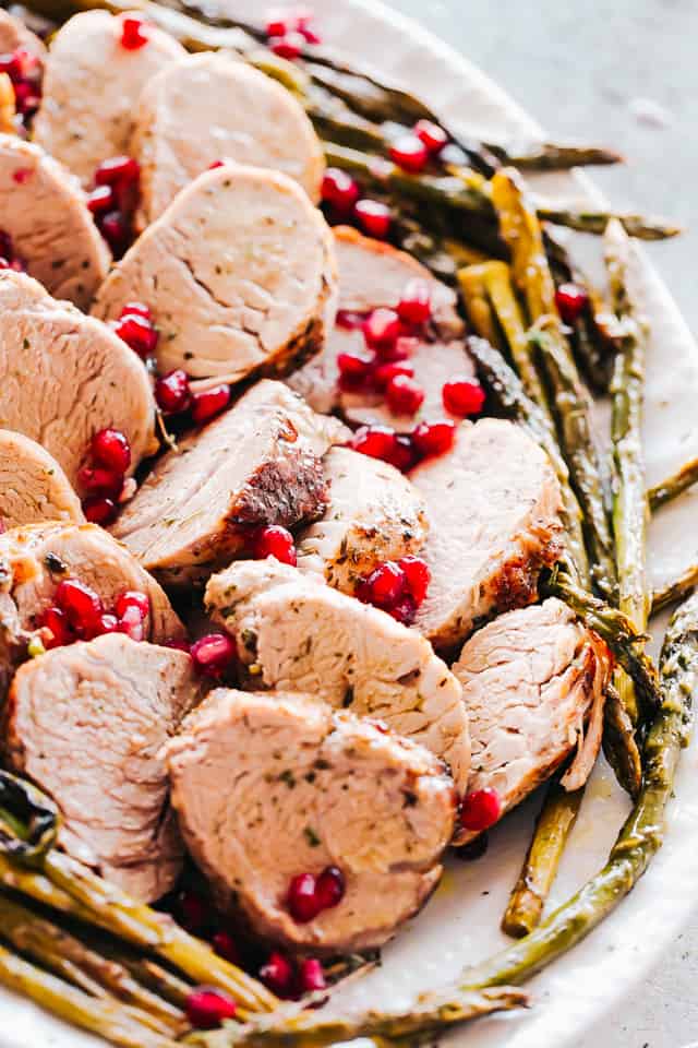 A white serving platter containing roasted pork tenderloin and asparagus garnished with pomegranate seeds