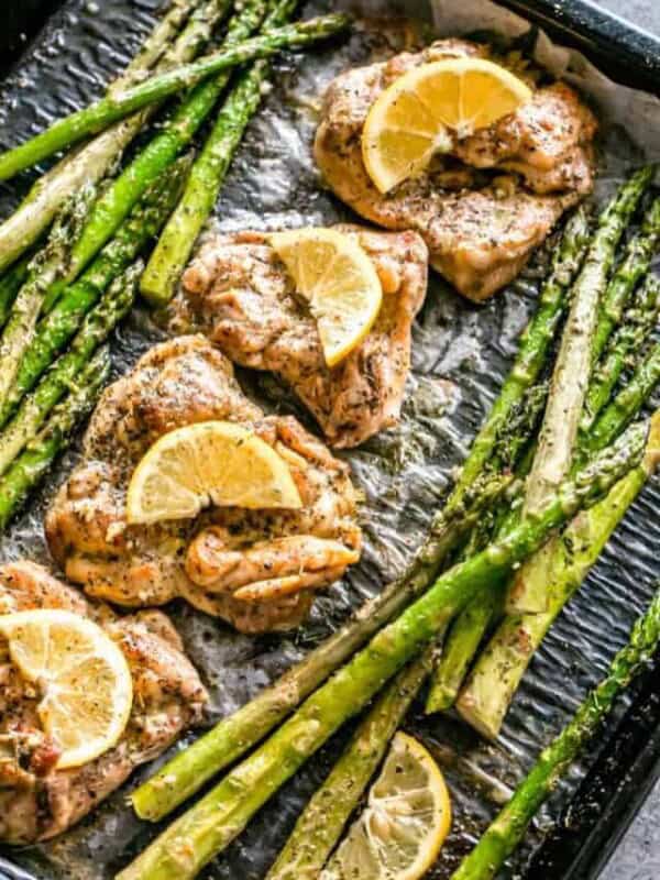 Overhead view of lemon garlic chicken thighs garnished with lemon slices, next to asparagus on a baking pan.