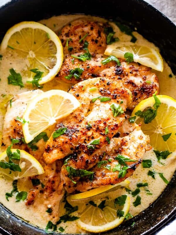 Overhead view of lemon parmesan chicken in a skillet with creamy sauce, topped with lemon slices and fresh herbs.