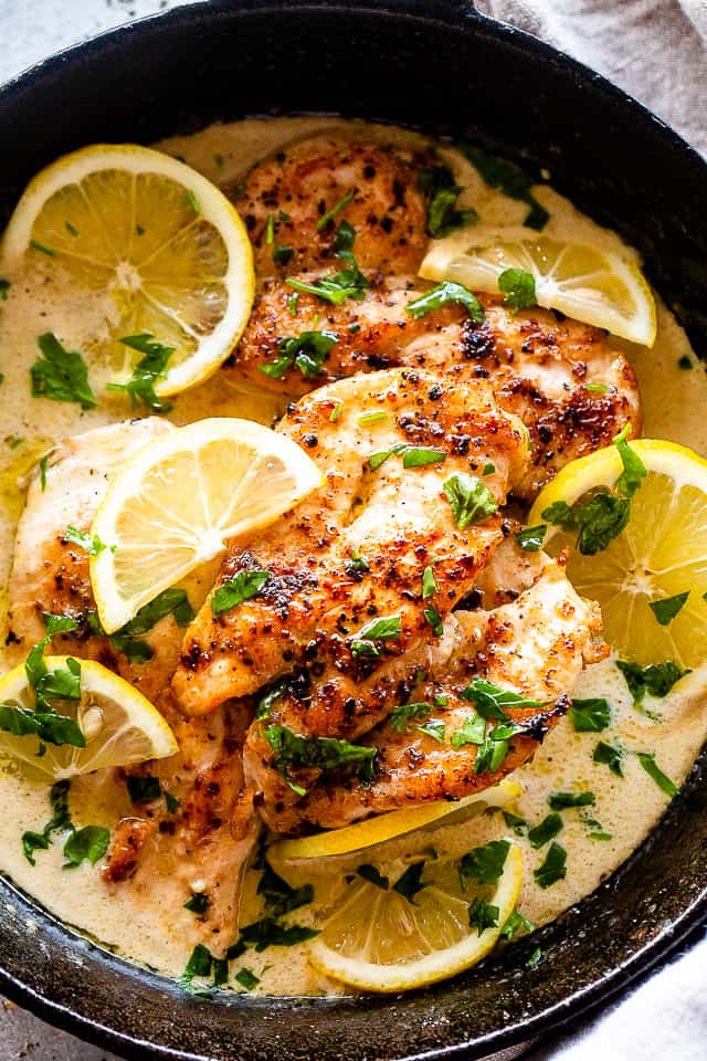 Overhead view of lemon parmesan chicken in a skillet with creamy sauce, topped with lemon slices and fresh herbs.