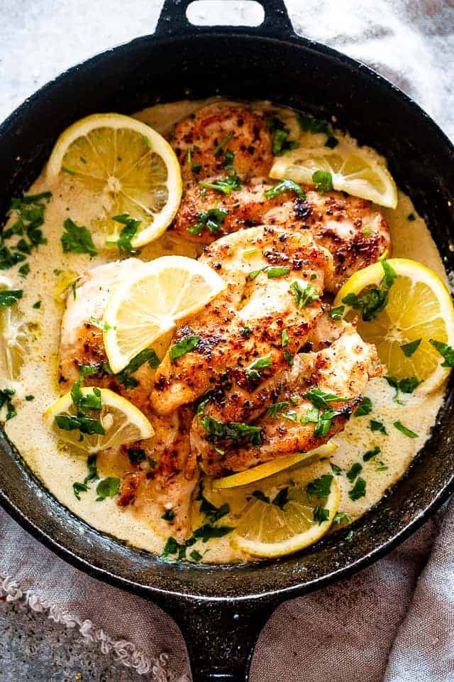 Overhead view of lemon parmesan chicken in a skillet with creamy sauce, topped with lemon slices and fresh herbs.