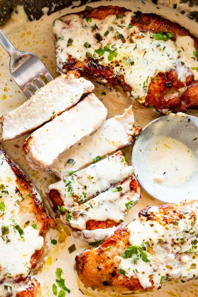 A pork chop cut into slices, with a fork running through two slices, next to three whole pork chops, all covered in a cream sauce and herbs. 