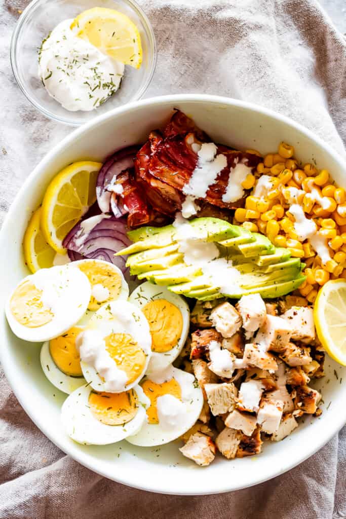 Overhead shot of a bowl with sliced hard boiled eggs, diced cooked chicken, slices of avocado, crumbled bacon, corn kernels, and two slices of lemon.