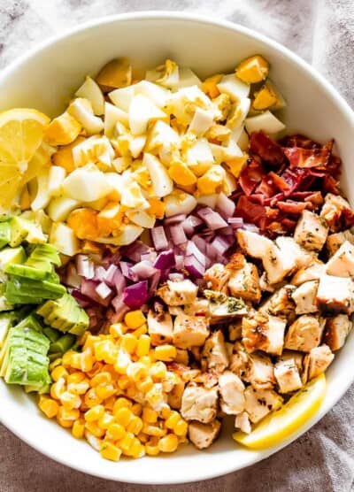 overhead shot of a white salad bowl with chopped hard boiled eggs, sliced avocados, diced onions, diced cooked chicken, crumbled bacon, and corn kernels.