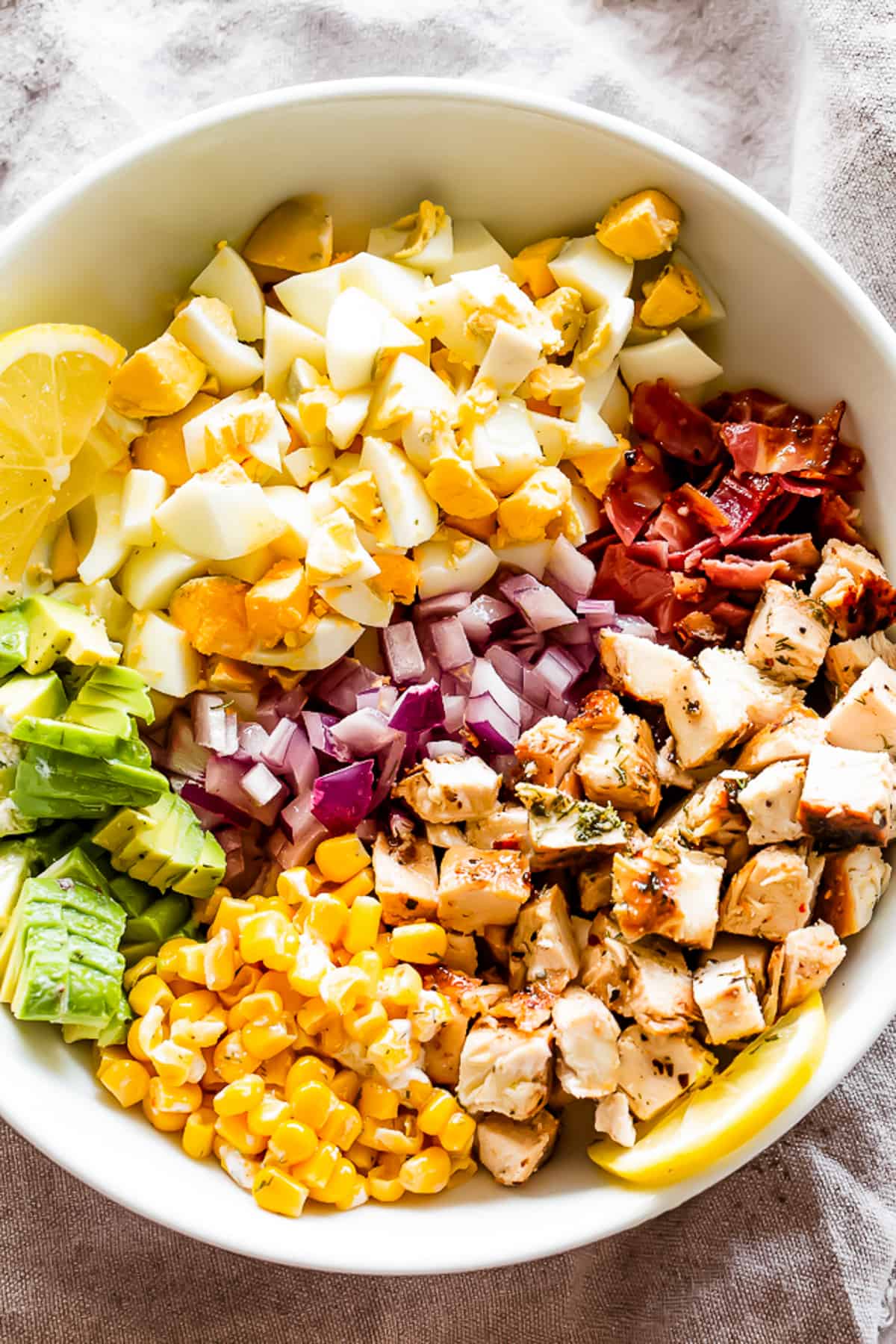 overhead shot of a white salad bowl with chopped hard boiled eggs, sliced avocados, diced onions, diced cooked chicken, crumbled bacon, and corn kernels.