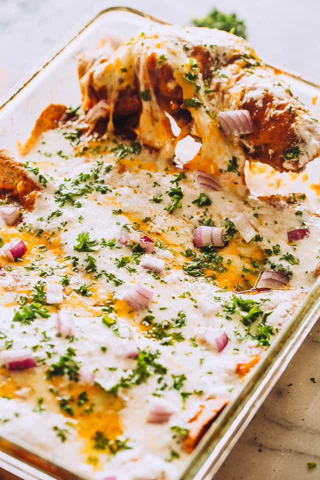 A serving being lifted from a beef enchiladas topped with cheese and sour cream in a baking dish.