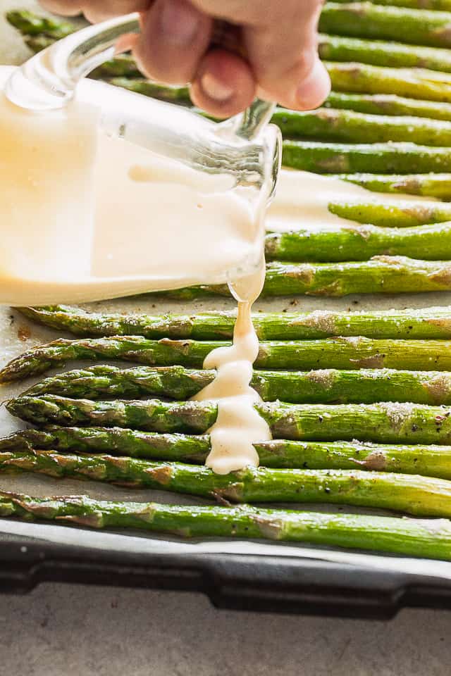 Pouring sauce over roasted asparagus on a baking sheet.