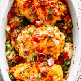 three baked chicken breasts in a white baking dish, and topped with melted cheese, crumbled bacon, and sliced green onions.