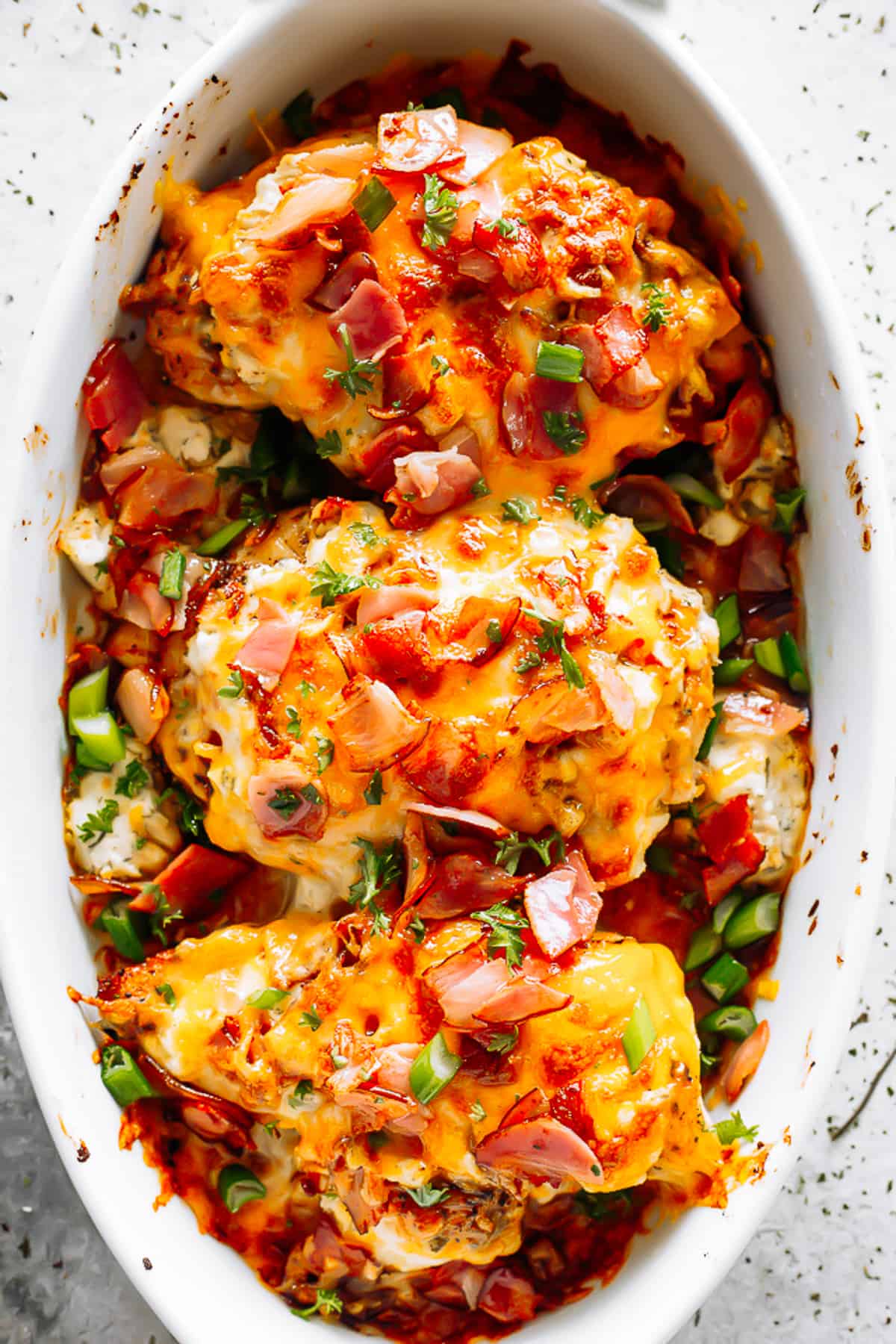 three baked chicken breasts in a white baking dish, and topped with melted cheese, crumbled bacon, and sliced green onions.
