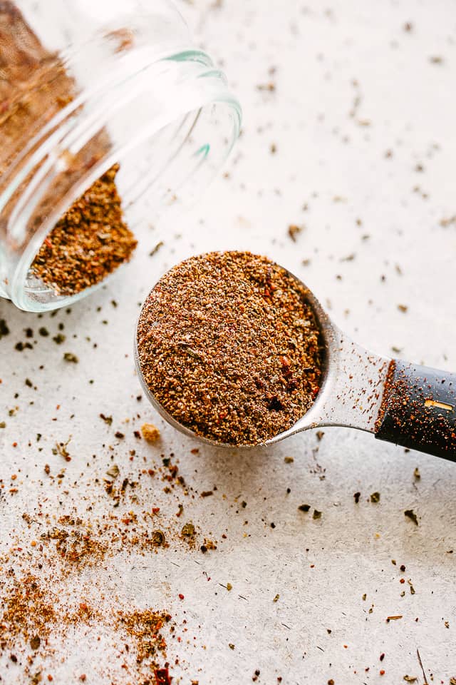 A spoon full of Homemade Taco Seasoning next to a jar of seasoning tipped on its side.