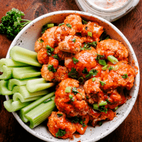 Cauliflower florets topped with hot sauce and served in a bowl with celery sticks. Bleu cheese dressing is placed at the top right of the bowl.
