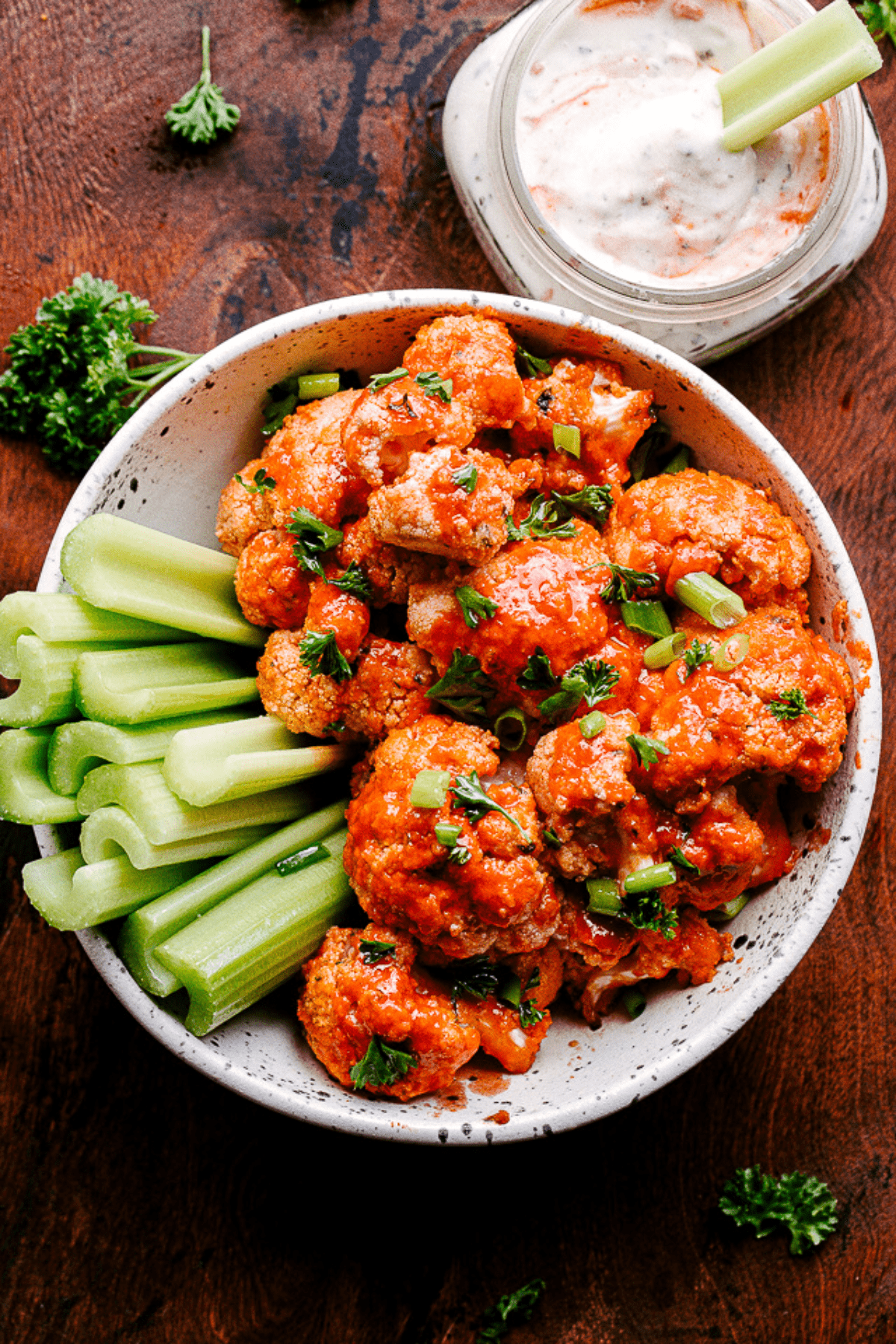 Cauliflower florets topped with hot sauce and served in a bowl with celery sticks. Bleu cheese dressing is placed at the top right of the bowl.