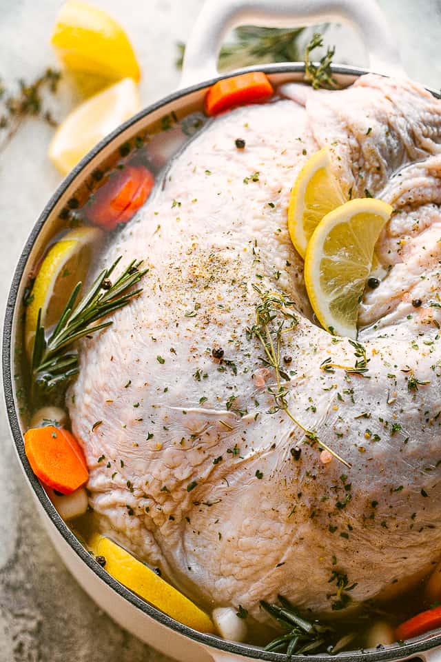 A whole turkey submerged in a brine with herbs, garlic, carrots, and lemon slices.