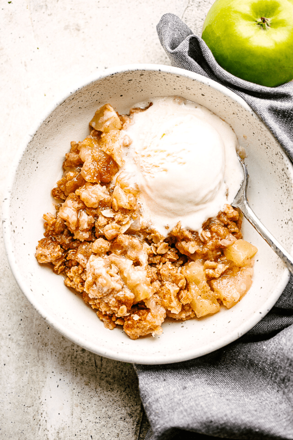 overhead shot of Apple Crumble served in a bowl with vanilla ice cream, and a green apple placed to the right side of the bowl.