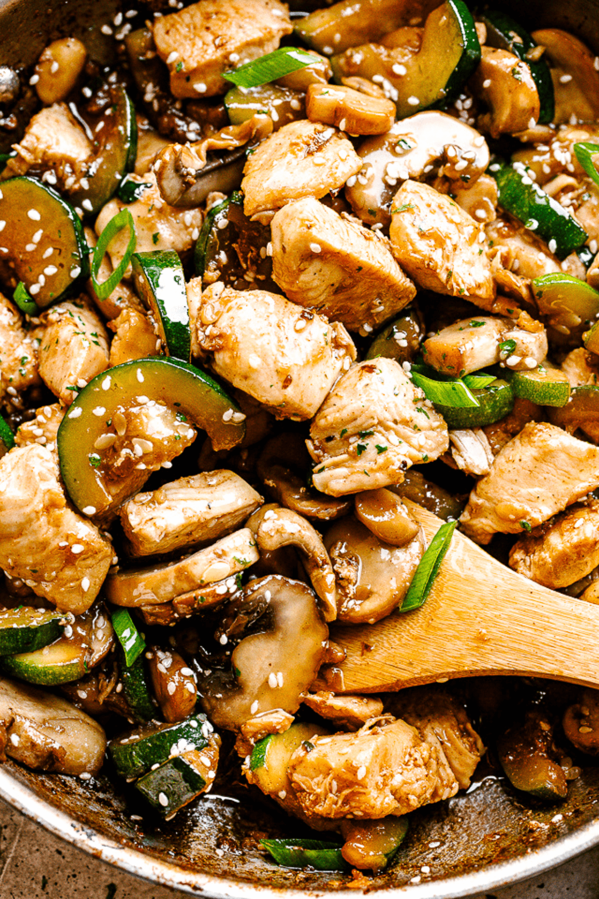 close up photo of wooden spoon stirring through chicken stir fry with zucchini and mushrooms.