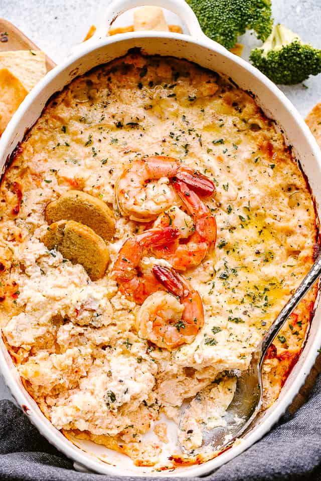 cream cheese shrimp dip in a baking dish, topped with additional shrimp, bagel chips, and a sprinkling of herbs.