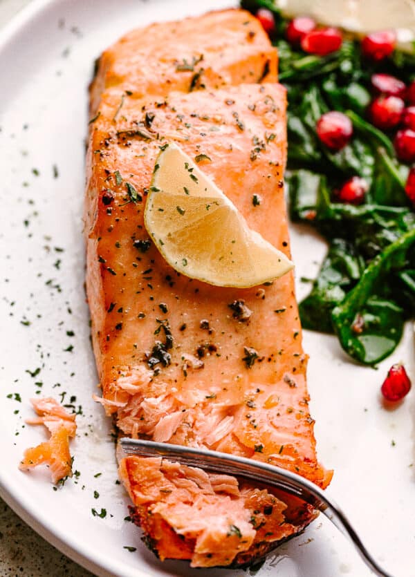 Salmon fillet garnished with lemon, on a plate with greens and pomegranate arils.