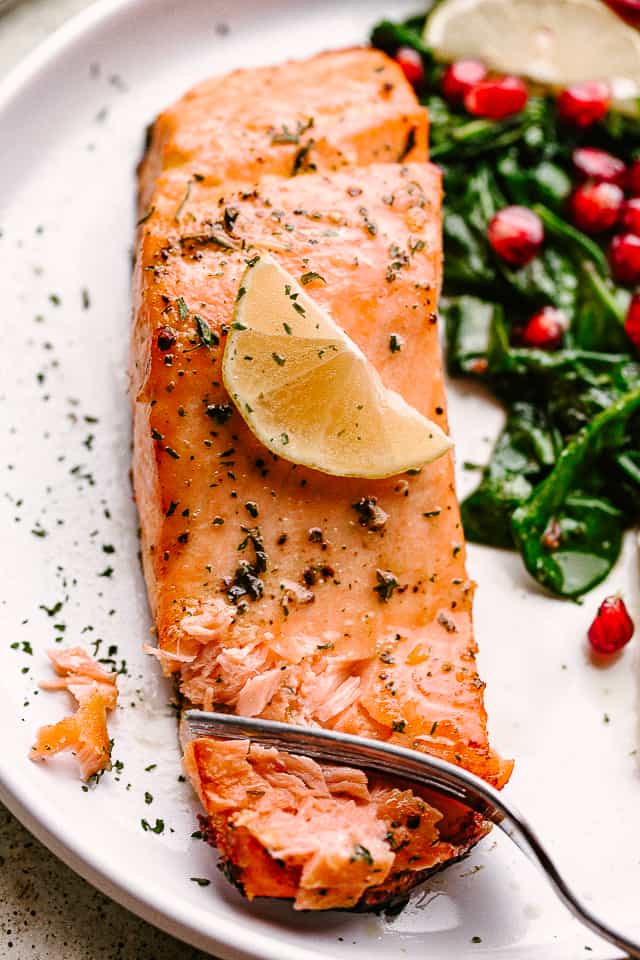 Salmon fillet garnished with lemon, on a plate with greens and pomegranate arils.