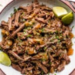 Pulled beef barbacoa served in a large bowl with lime wedges.