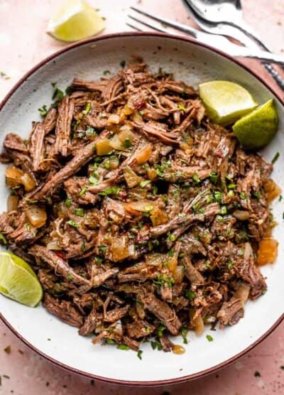 Pulled beef barbacoa served in a large bowl with lime wedges.