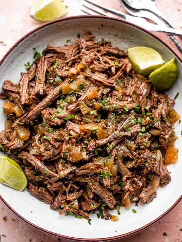 Pulled beef barbacoa served in a large bowl with lime wedges.