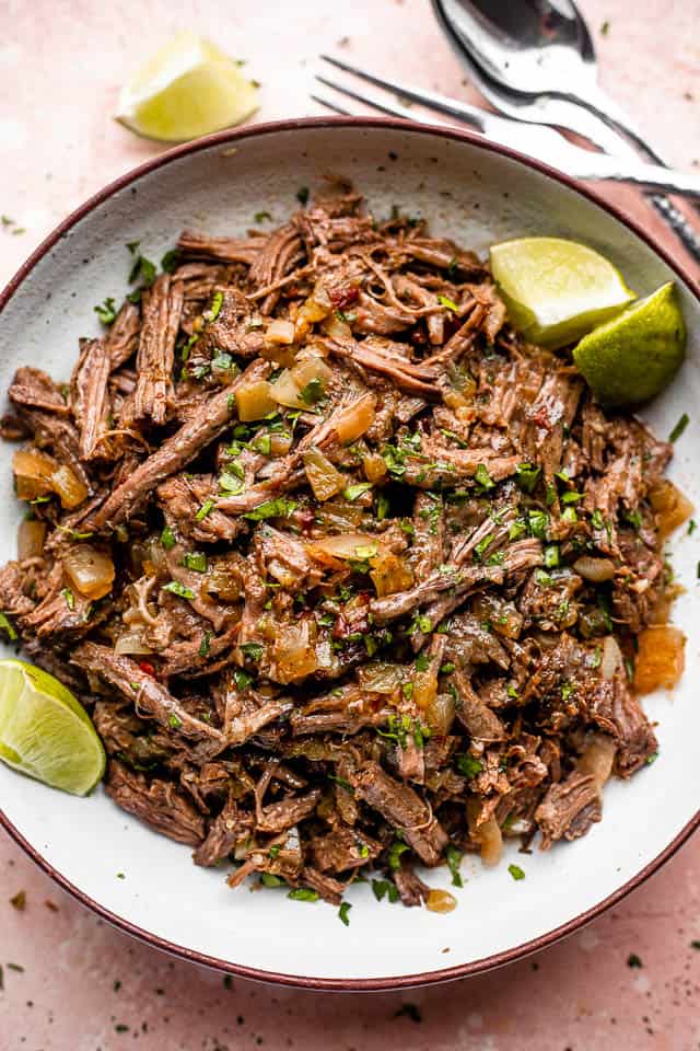 Pulled beef barbacoa served in a large bowl with lime wedges.