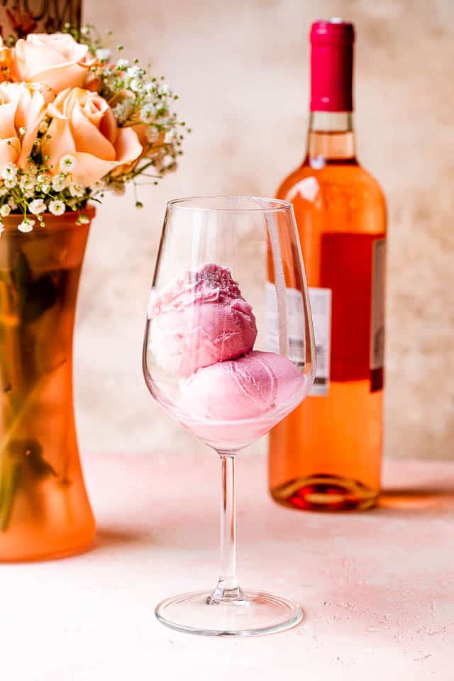 Two scoops of sorbet in a wine glass in front of a vase of roses and a bottle of rosé.
