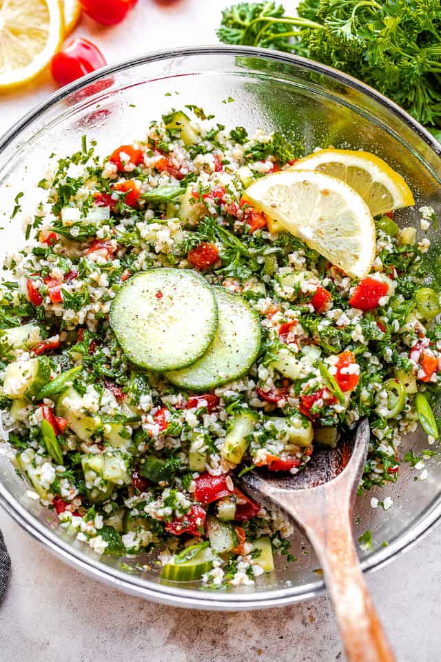 Tabouli Salad in a glass salad bowl with parsley, lemons, and tomatoes on the side.