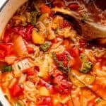 A wooden spoon stirring through a soup with cabbage, tomatoes, zucchini, and broccoli.