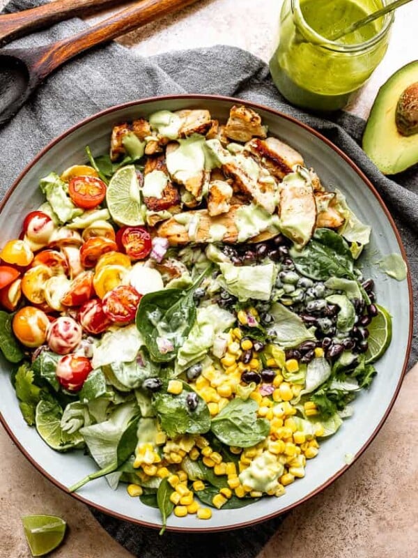 a jar of salad dressing and two wooden spoons set next to a salad bowl filled with chicken strips, corn, tomatoes, and black beans