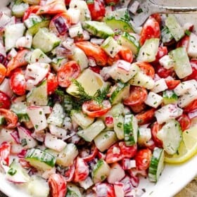 top view of a silver spoon inside a white salad bowl filled with chopped tomatoes, cucumbers, radishes, herbs, and tossed with a creamy dressing