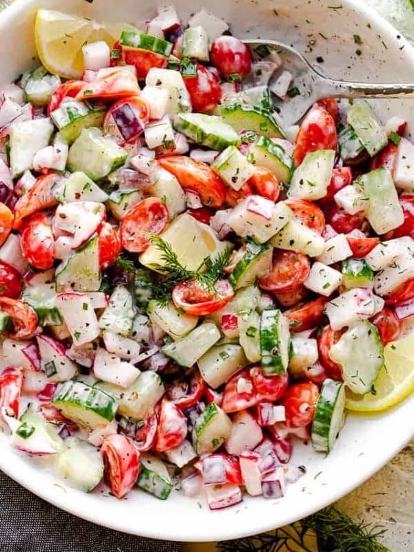 top view of a silver spoon inside a white salad bowl filled with chopped tomatoes, cucumbers, radishes, herbs, and tossed with a creamy dressing