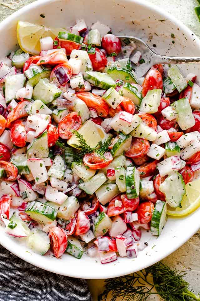 top view of a silver spoon inside a white salad bowl filled with chopped tomatoes, cucumbers, radishes, herbs, and tossed with a creamy dressing