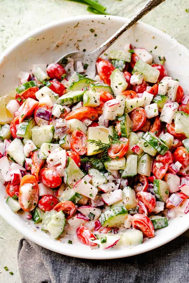 large silver spoon inside a white bowl with chopped cucumbers, chopped tomatoes, and chopped radishes