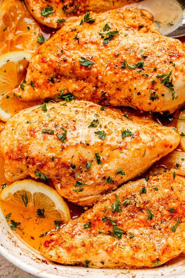 close up above view shot of baked chicken breasts in a white baking dish sprinkled with parsley and garnished with lemons