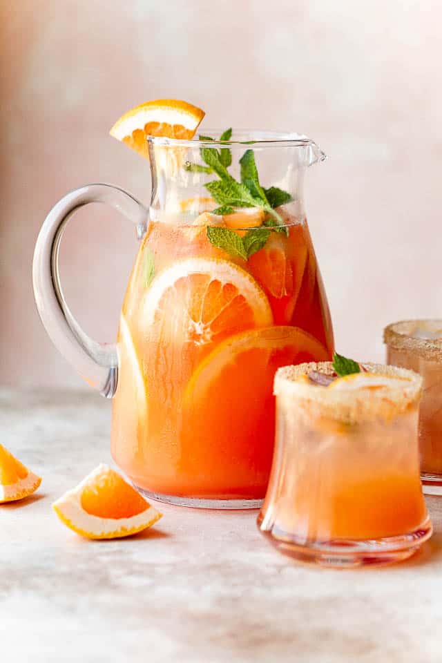 large glass pitcher filled with grapefruit rum cocktail, mint leaves, and two drinking glasses next to the pitcher