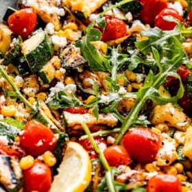 side shot of a blue salad bowl filled with grilled corn, tomatoes, zucchini, goat cheese, lemon slices, and arugula on top
