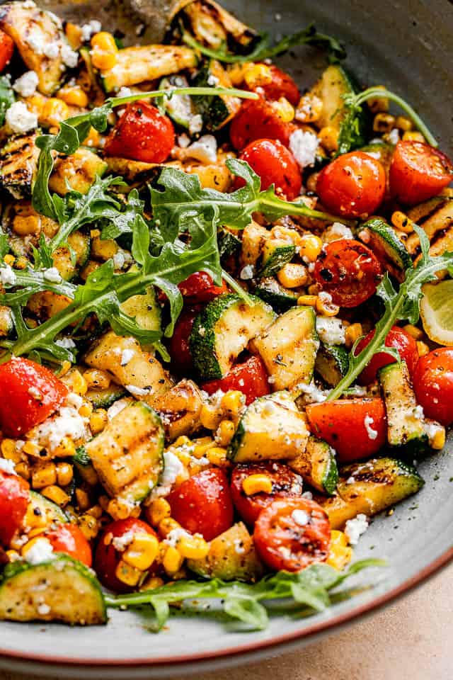 close up shot of a salad bowl filled with corn kernels, tomatoes, zucchini, cheese, and arugula on top