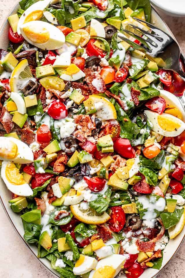 top view shot of boiled egg wedges, lemon slices, chopped avocados, tomatoes, and bacon set on top of baby spinach leaves and salad greens