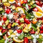 top view shot of boiled egg wedges, lemon slices, chopped avocados, tomatoes, and bacon set on top of baby spinach leaves and salad greens