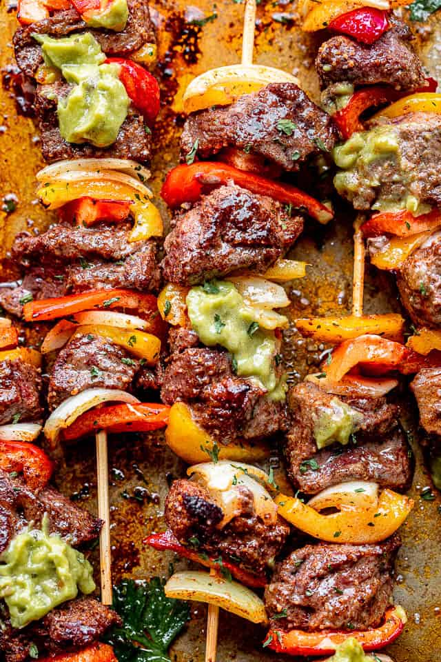 close up shot of steak and vegetable kabobs set over a plate and topped with guacamole