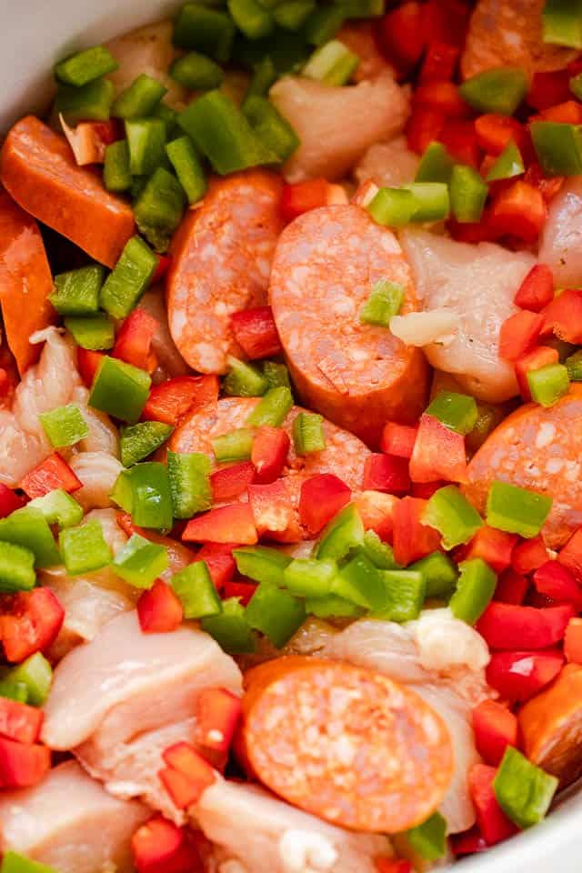 A slow cooker insert filled with diced green peppers, diced red peppers, chopped chicken, and sliced sausages.