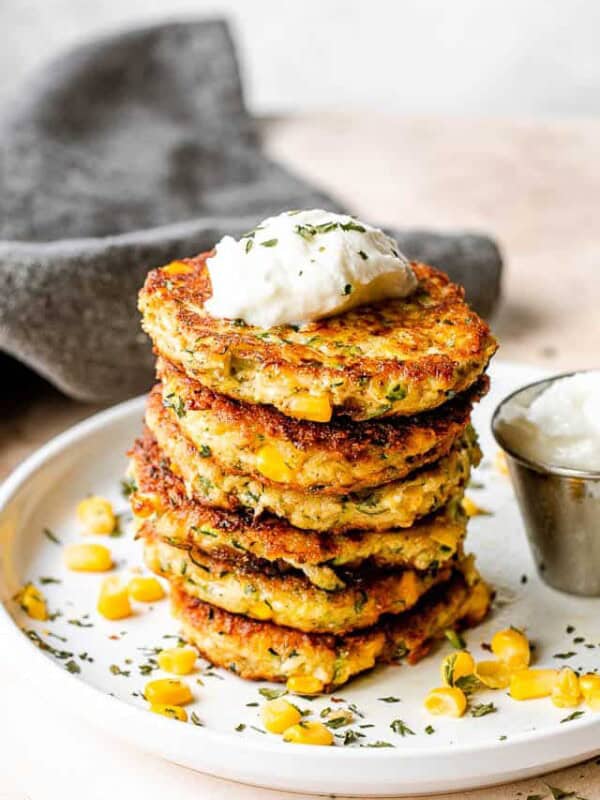 stack of zucchini corn fritters set on a white plate with yogurt dip next to it, and a gray linen napkin behind the plate