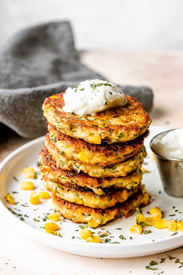 Stack of zucchini corn fritters set on a white plate with yogurt dip next to it and a gray linen napkin behind the plate.