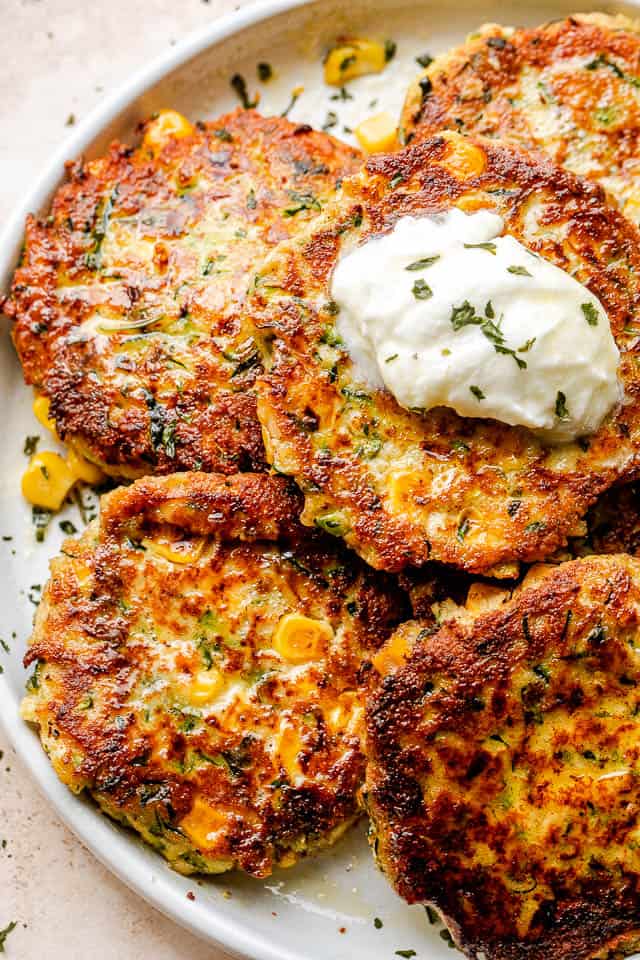 Arranged corn zucchini fritters on a plate and topped with a dollop of sour cream and a sprinkle of dried herbs.