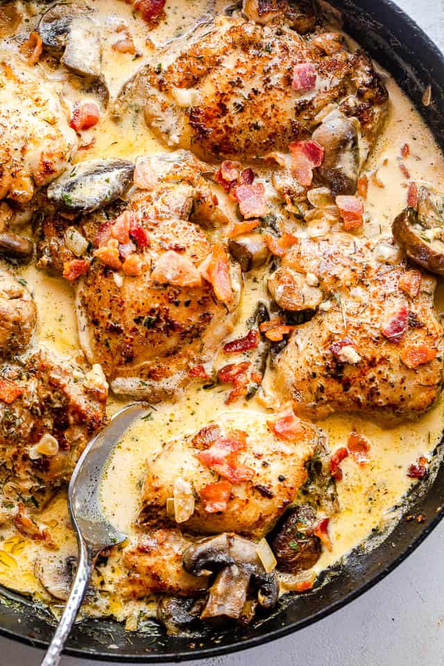 Close-up shot of a spoon inside a black skillet filled with boneless chicken thighs cooking in a cream sauce with mushrooms and bacon