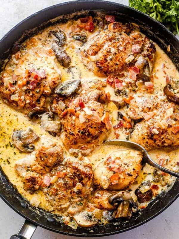 top shot of a black skillet with boneless chicken thighs cooking in a cream sauce with mushrooms and bacon