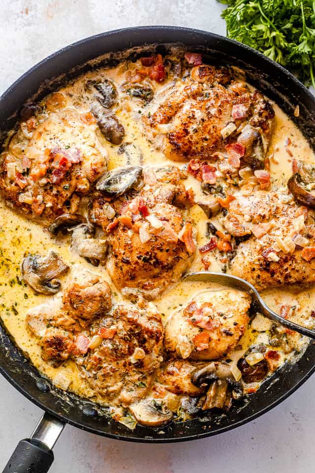 top shot of a black skillet with boneless chicken thighs cooking in a cream sauce with mushrooms and bacon